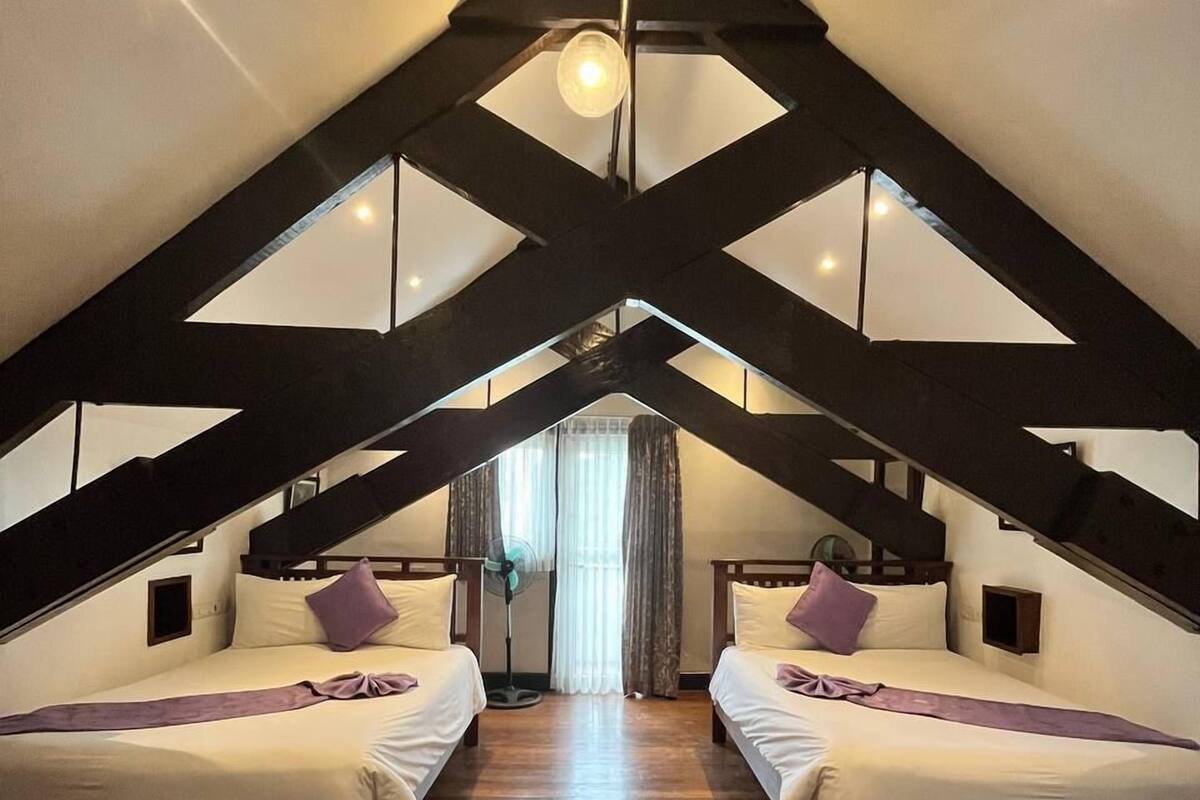 A room with two beds, wooden flooring, a ceiling fan, and a window with curtains.