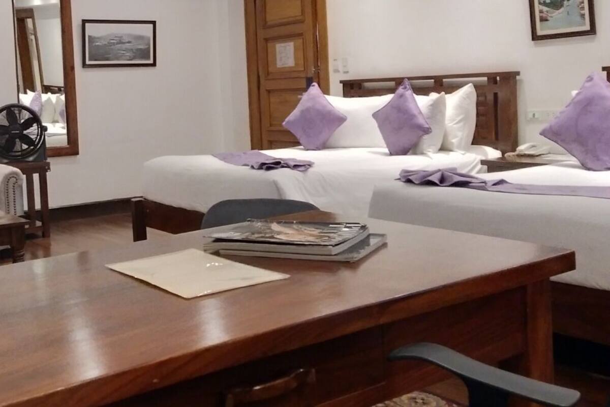 A hotel room with a wooden desk, a sofa, a bed with white and purple pillows, a mirror, and a framed picture on the wall.