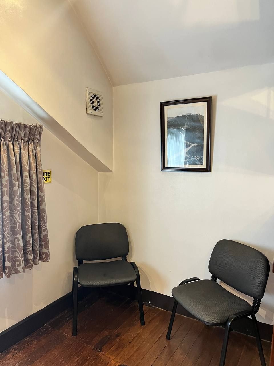 Habitación de esquina con dos sillas negras, un cuadro en la pared, ventana con cortinas y señal de salida visible.