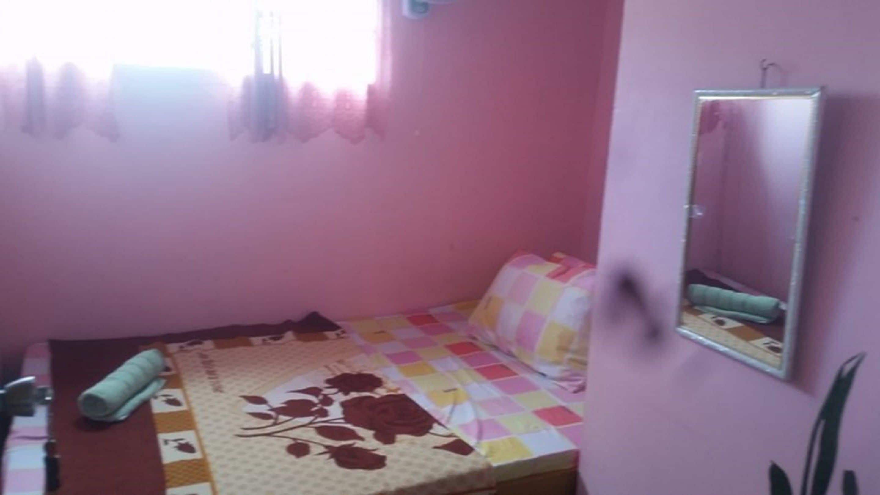 Una habitación pequeña con paredes rosadas, una cama, un espejo y una ventana con cortinas.