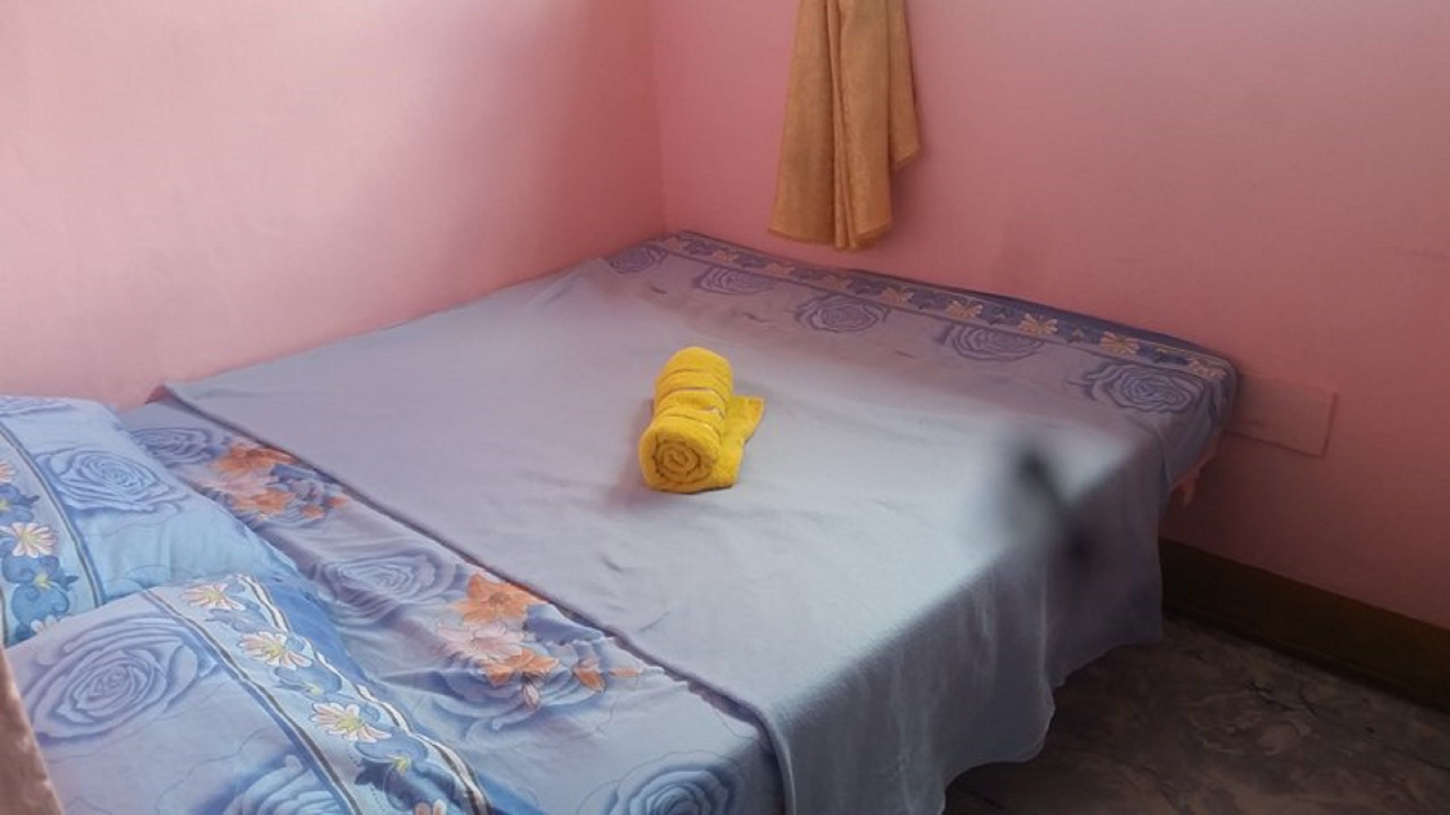 Una habitación pequeña con cama individual, pared rosa, colcha estampada y una toalla enrollada sobre la cama.
