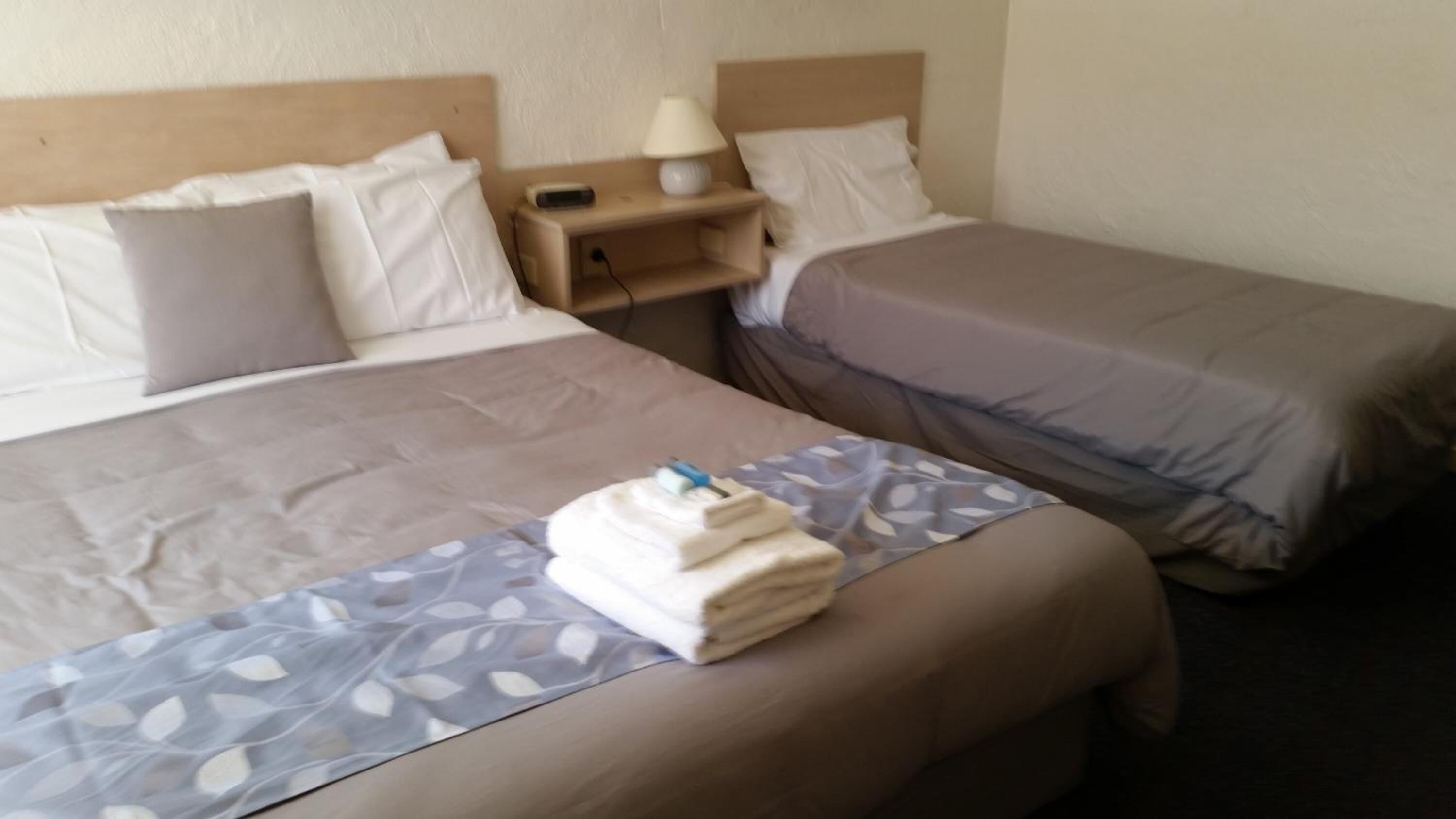 A hotel room with two beds, a nightstand with a lamp, and a folded towel on the bed.