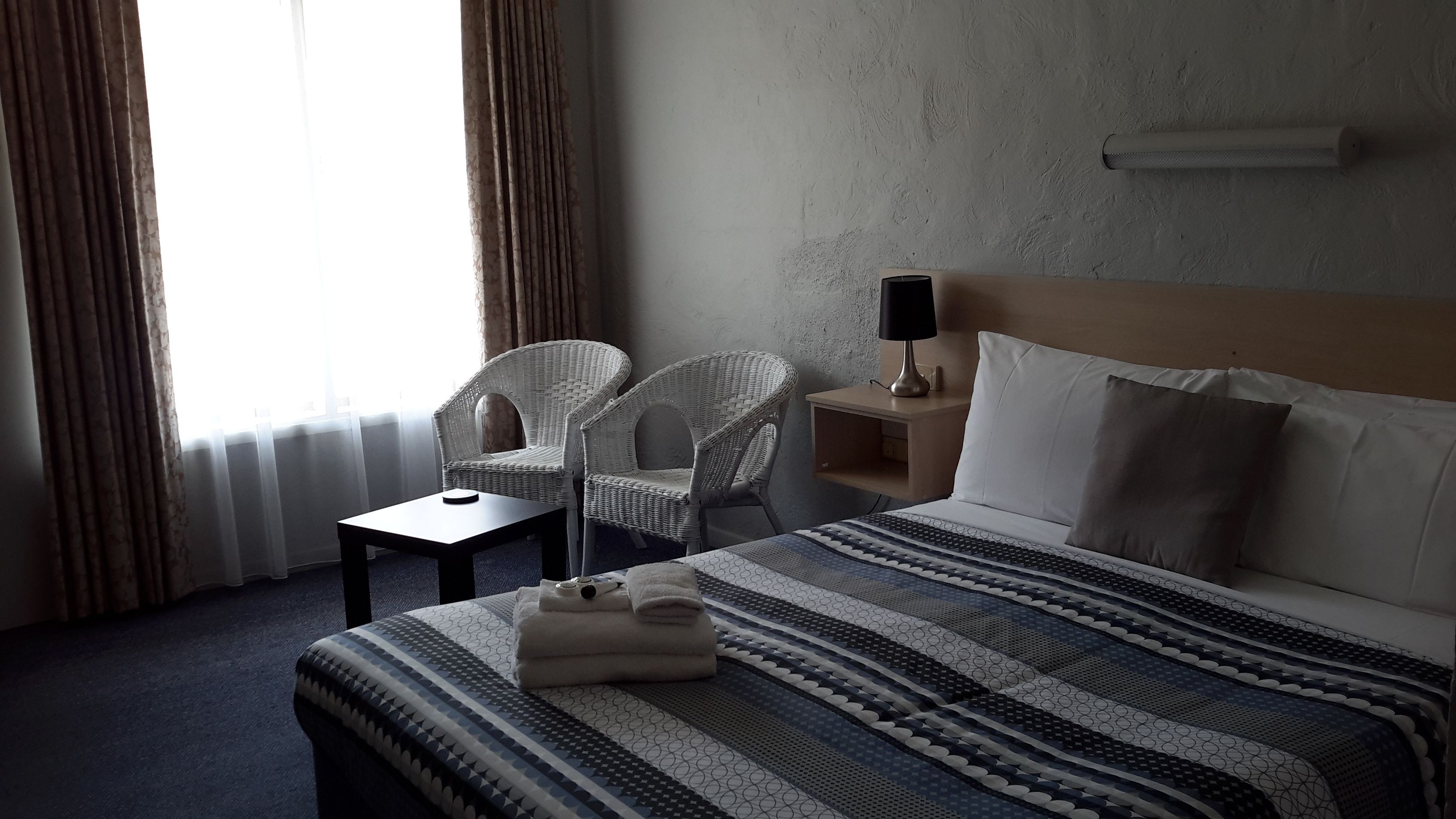 Una habitación de hotel con una cama grande, dos sillones de mimbre, una mesita, una lámpara de noche y vistas al exterior a través de cortinas translúcidas.