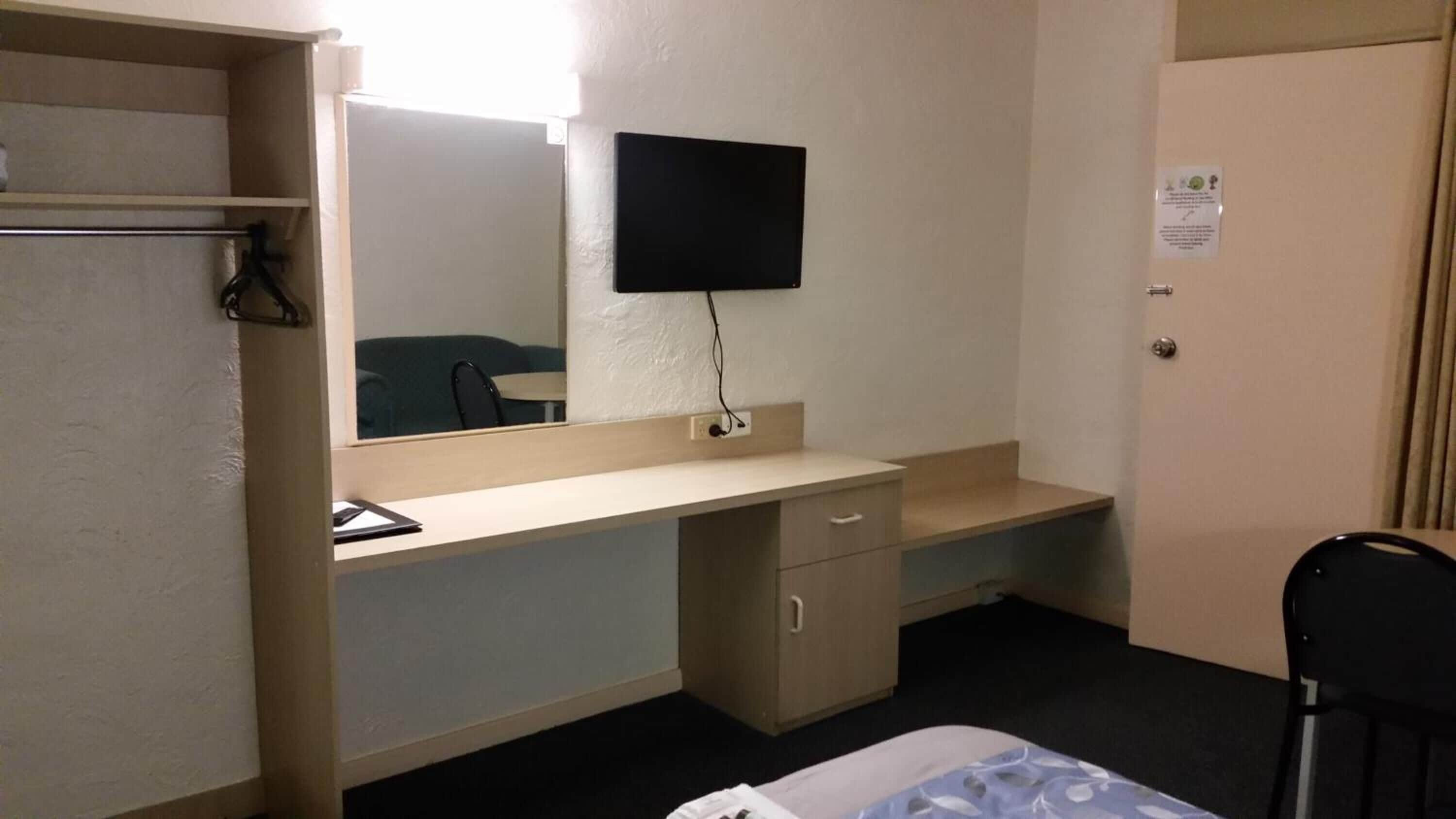 A hotel room with a bed, a desk with a chair, a mirror, a TV mounted on the wall, and a door.