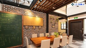 Une salle de conférence avec une table en bois, des chaises blanches, un tableau noir et un mur en briques.