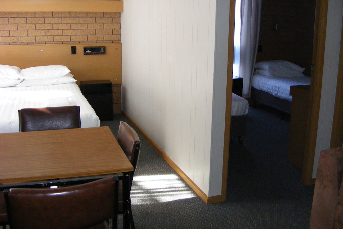 Ett hotellrum med en enkel säng, ett träbord och en stol. Det finns en tegelvägg och en vit dörr som leder till ett annat rum.