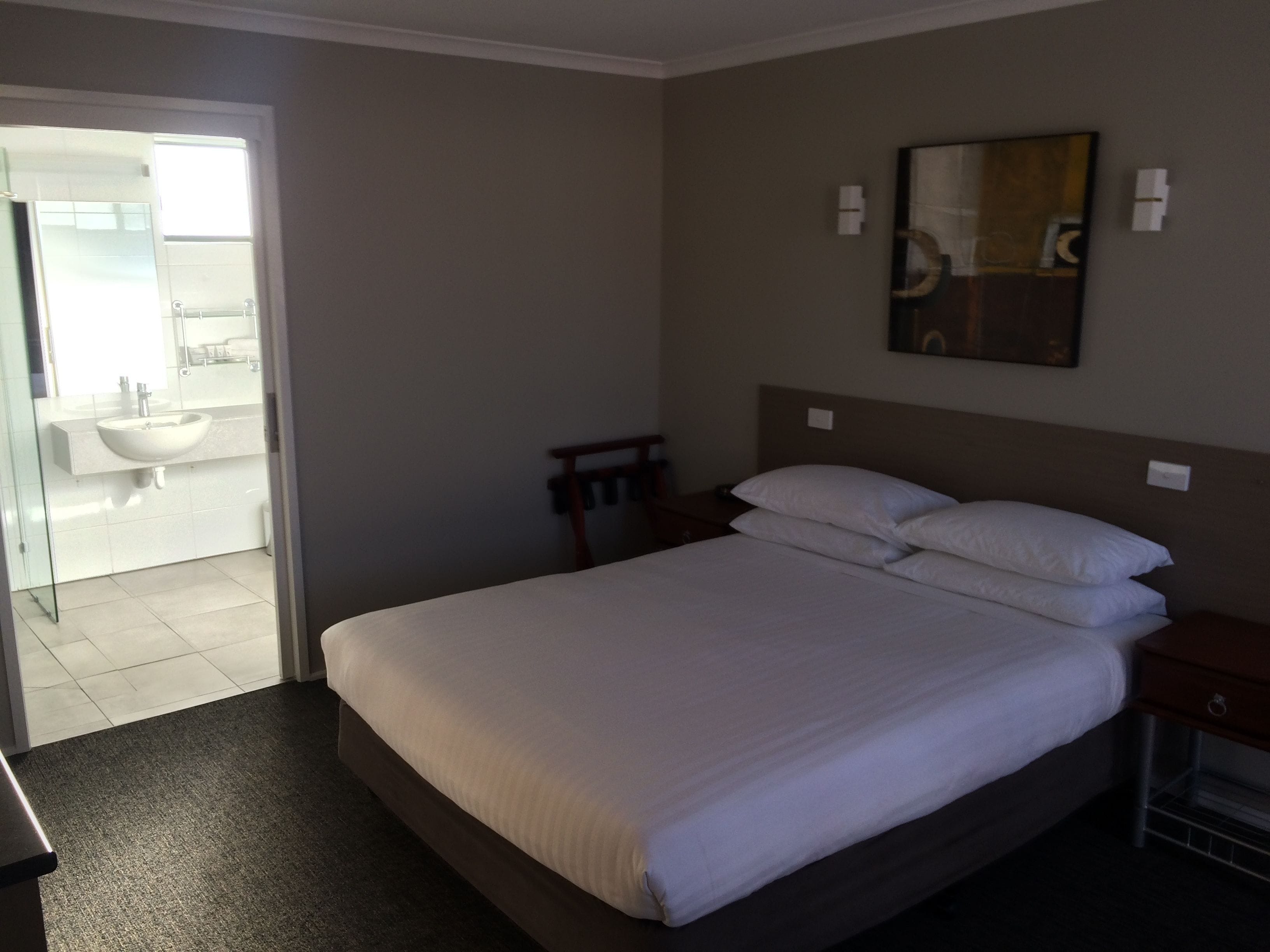 Ein Hotelzimmer mit einem großen Bett, einem Nachttisch, einem Schreibtisch, einem Bild an der Wand und einem sichtbaren Badezimmer durch eine offene Tür.