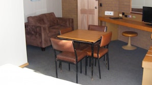 Een knus vertrek met een bruine leren bank, een houten eettafel met vier stoelen, een klein barretje met een kruk en een deur met lichtschakelaar.