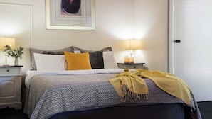 A neatly made bed with a patterned bedspread, a yellow throw blanket, and multiple pillows. There is a bedside table with a lamp and a vase with flowers. - Mahala 1 (Daylesford)