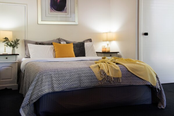 A neatly made bed with a patterned bedspread, a yellow throw blanket, and multiple pillows. There is a bedside table with a lamp and a vase with flowers.