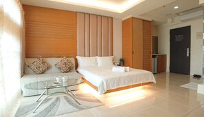Quarto de hotel moderno com uma cama, um sofá, uma mesa de centro de vidro e uma pequena cozinha.