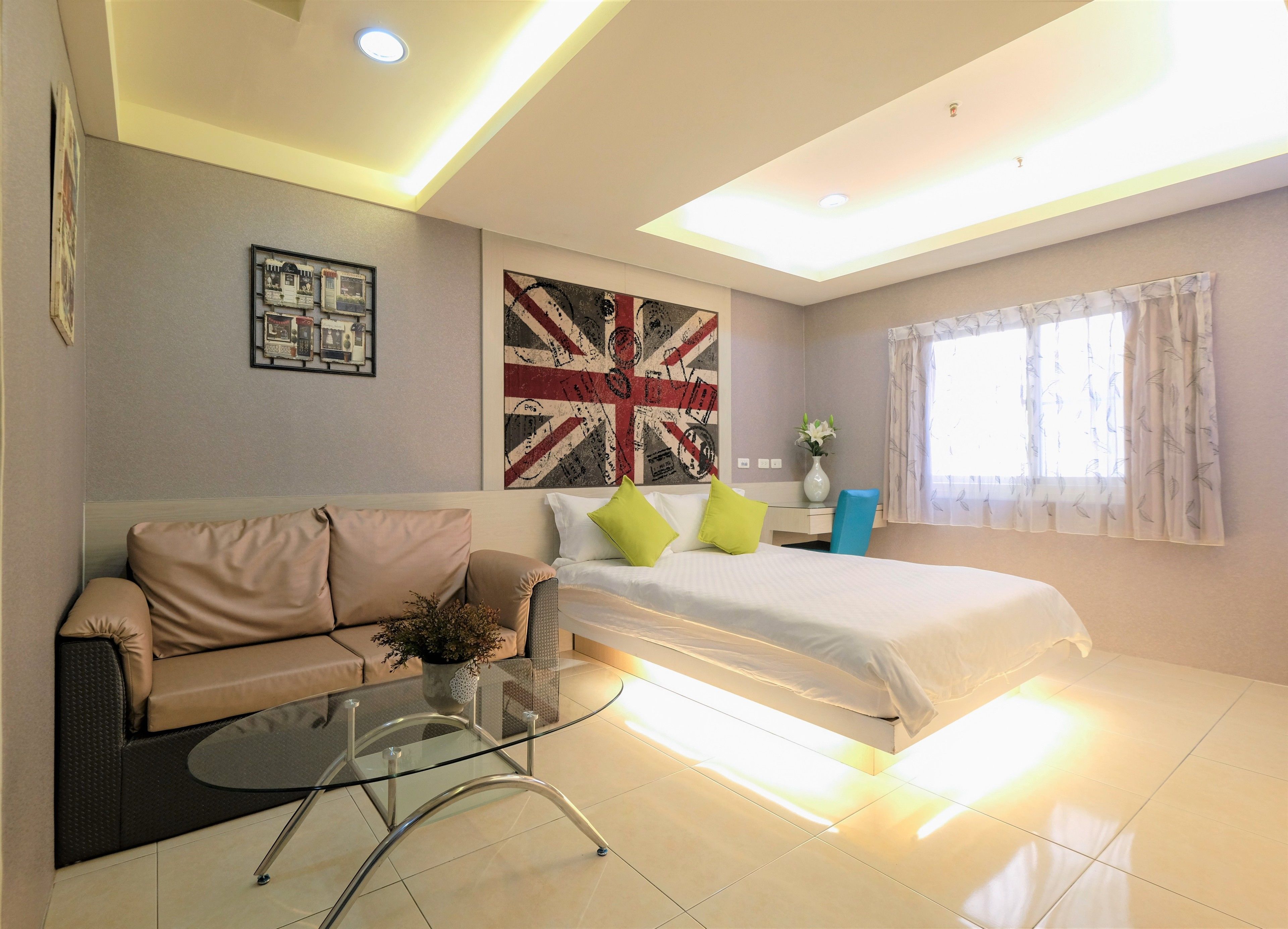 A modern hotel room with a bed, a sofa, a glass coffee table, and a wall with a Union Jack flag.