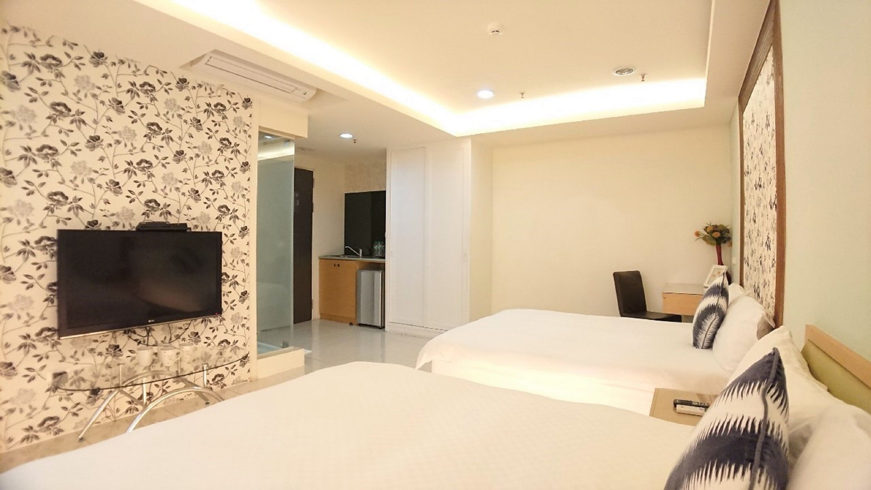 A hotel room with two beds, a TV mounted on the wall, a small kitchenette, and a dining area.