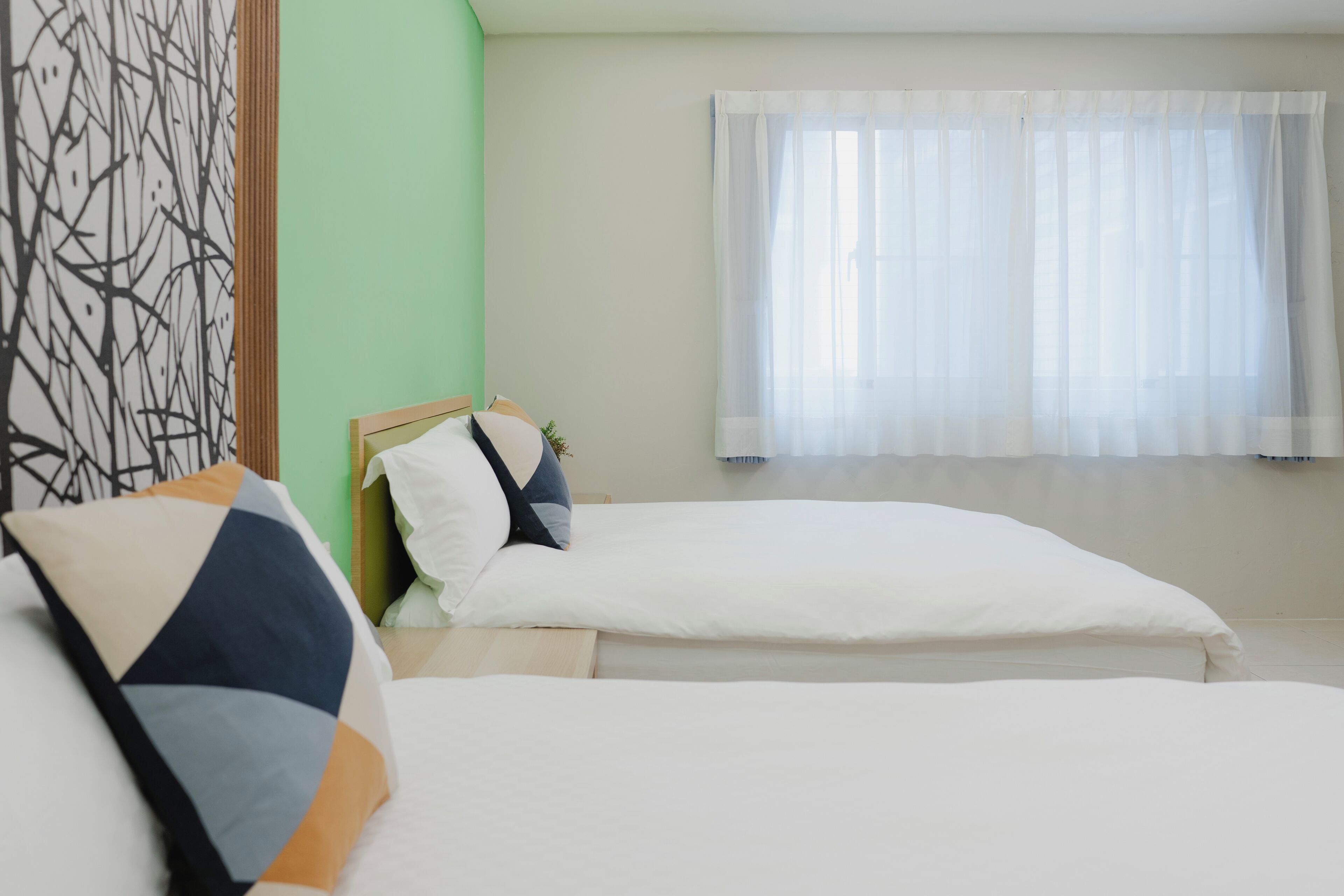 A bedroom with two beds, a green wall, and a window with sheer curtains.