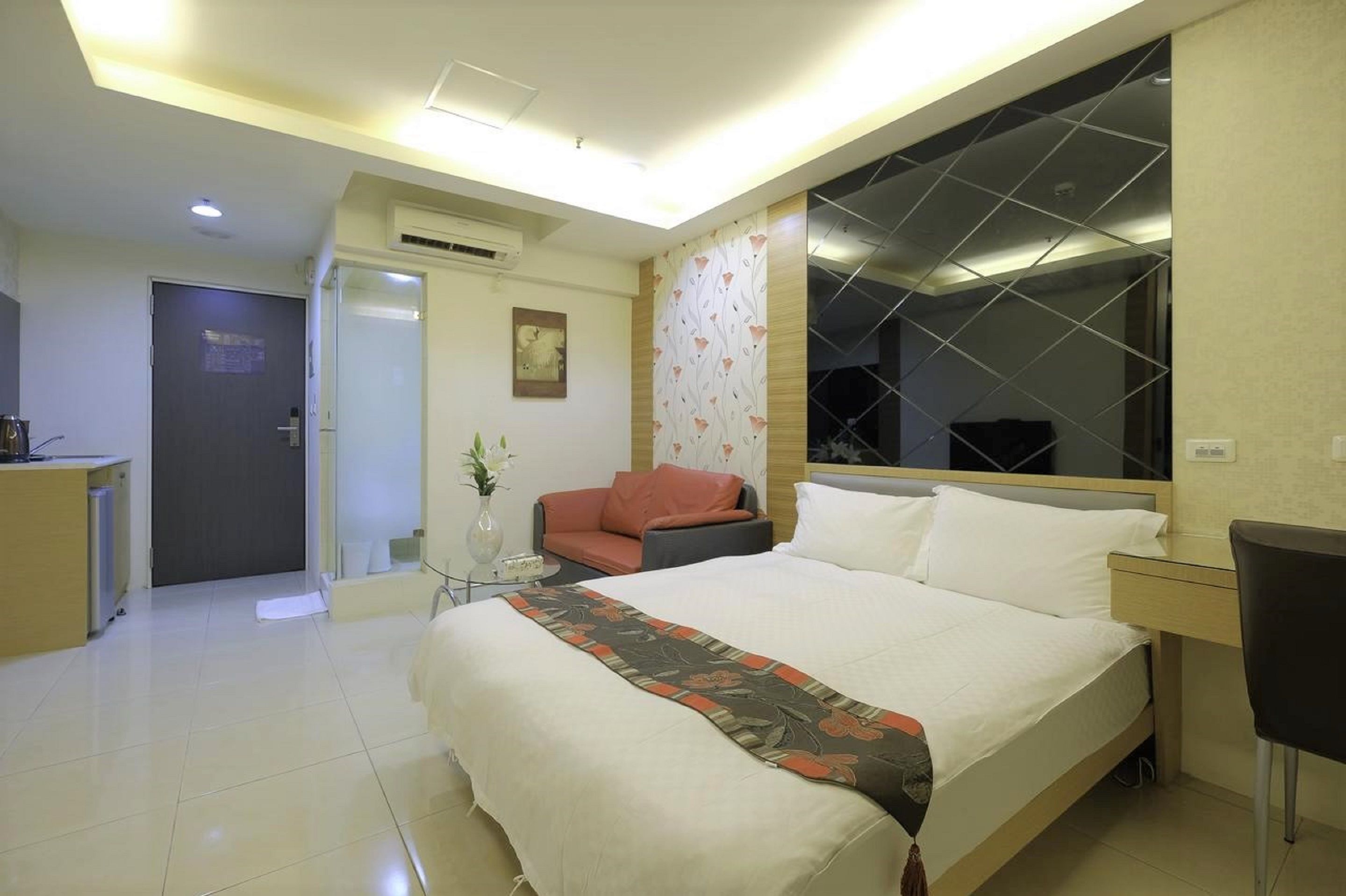 A hotel room with a bed, a desk, a chair, a sofa, and a small table with a vase.