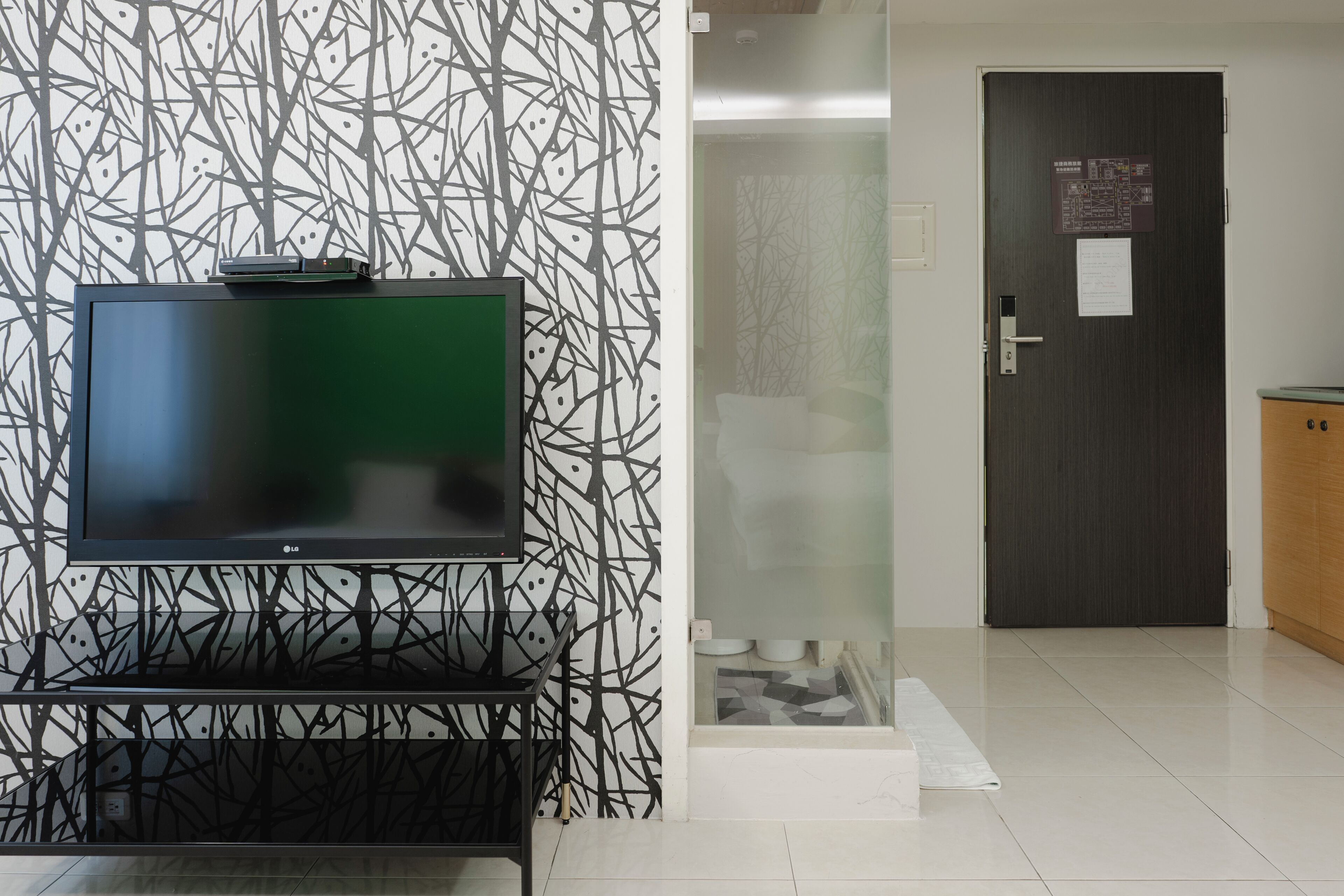 A modern hotel room with a flat-screen TV on a stand, a patterned wall, a door with a sign, and a tiled floor.