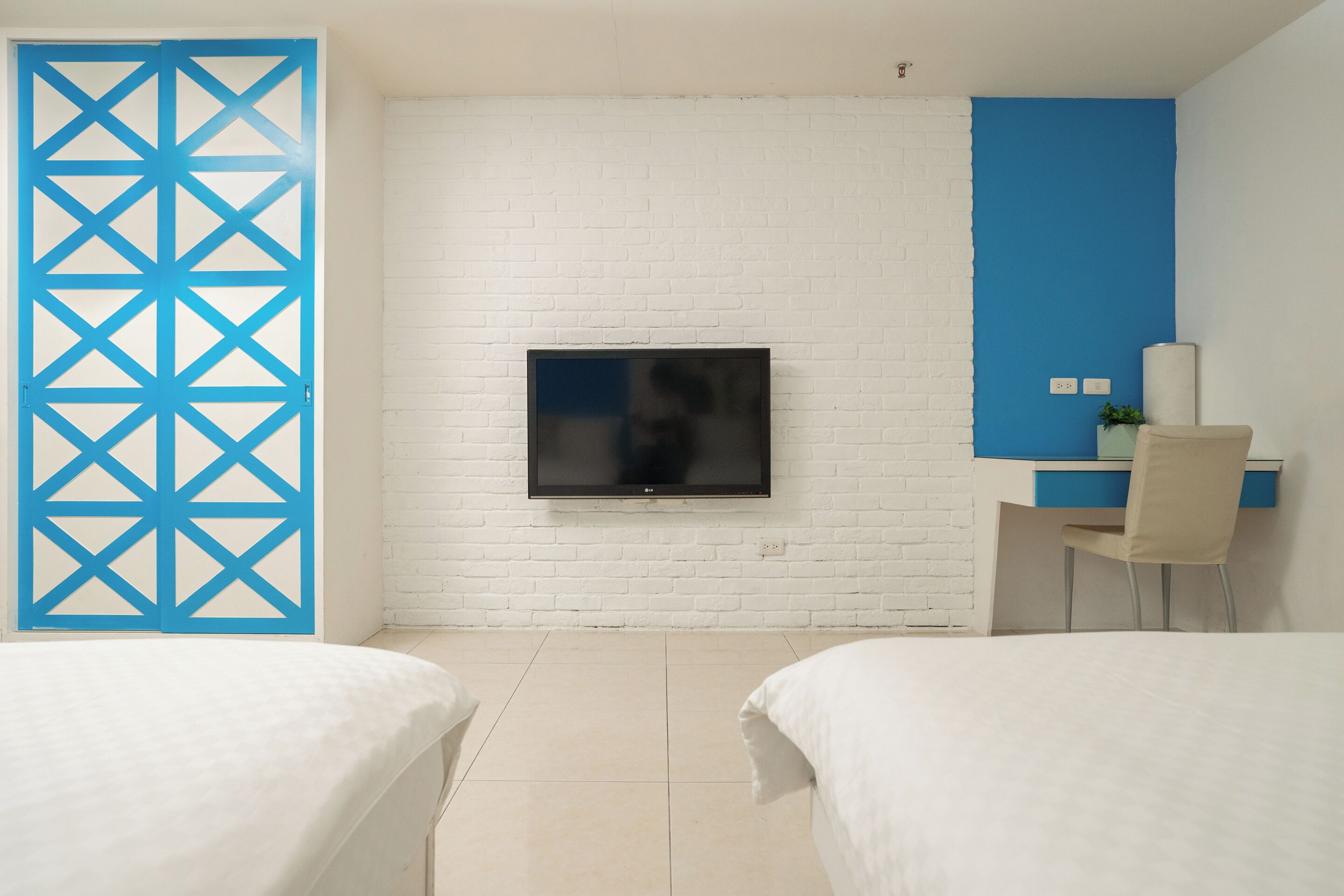 A hotel room with a white brick wall, a flat-screen TV mounted above a bed, a desk with a chair, and a blue partition.