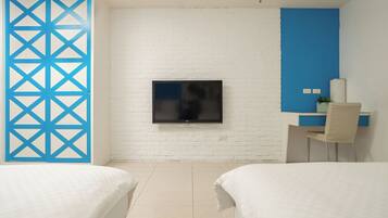 A hotel room with a white brick wall, a flat-screen TV mounted above a bed, a desk with a chair, and a blue partition.