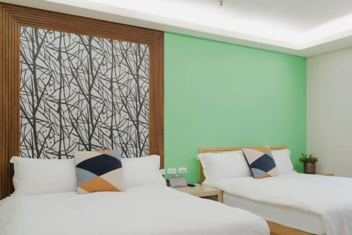 A hotel room with two beds, a green accent wall, and a decorative tree pattern on the left wall.