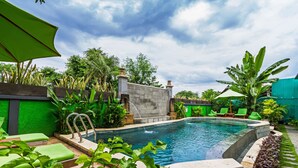 Una piscina con pared de piedra, sillas de descanso verdes y plantas tropicales.