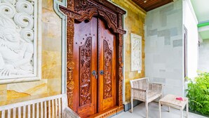 Una puerta de madera con intrincados grabados, una silla blanca y una pequeña mesa en un pasillo bien iluminado con suelo de baldosas.