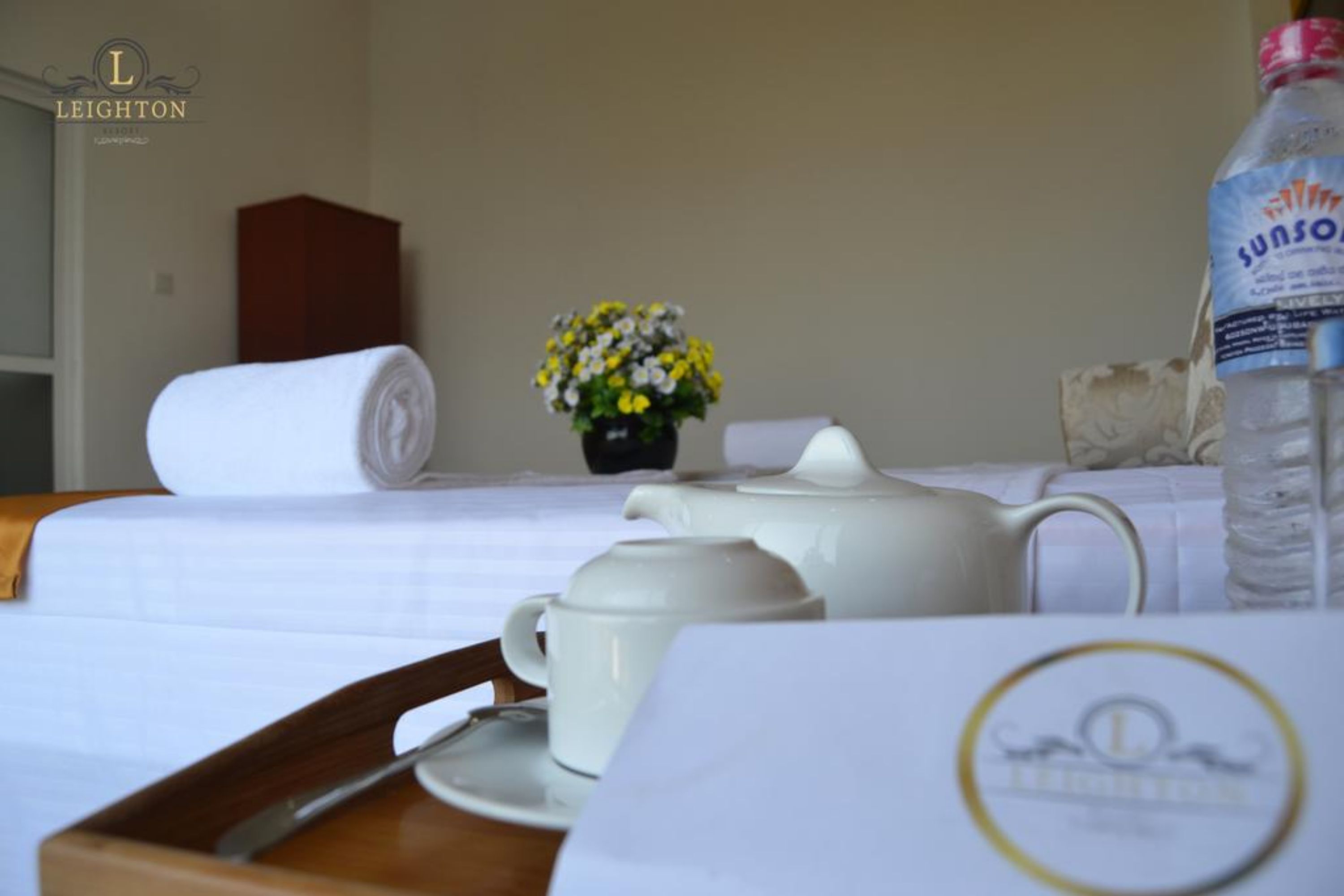 A neatly made bed with a rolled white towel, a white teapot, a white cup, and a bottle of water on a table.
