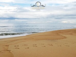 A beach with footprints leading to the water, and the Leighton Resort logo in the sky. - Leighton Resort (Negombo)