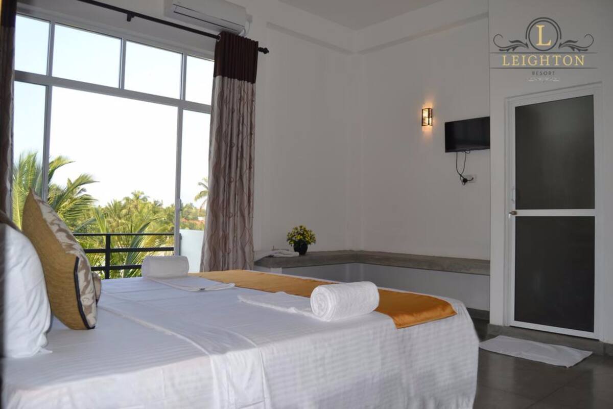 A hotel room with a bed, a window with curtains, a small table with a plant, and a wall-mounted TV.