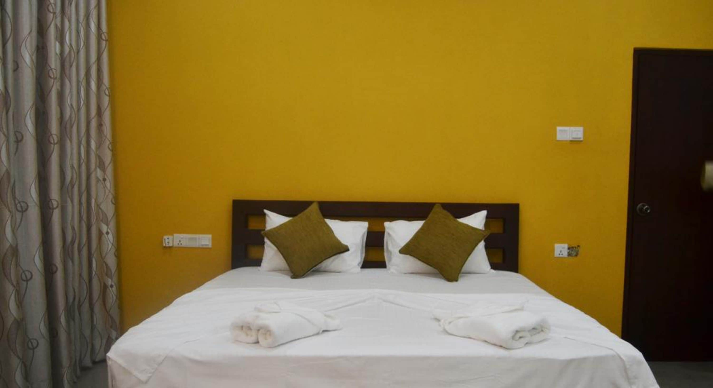 Ein Hotelzimmer mit gelber Wand, einem Holz-Kopfteil, zwei Kissen und einem ordentlich bezogenen Bett mit Handtüchern.
