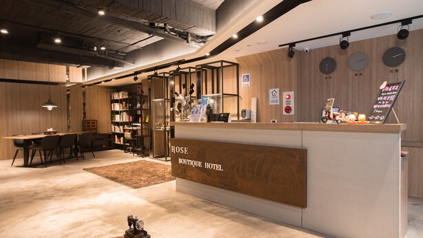 A reception area with a wooden reception desk labeled "ROSE BOUTIQUE HOTEL," a seating area with tables and chairs, and a bookshelf filled with books. - Royal Rose Hotel Shuangcheng Hall (Taipei)