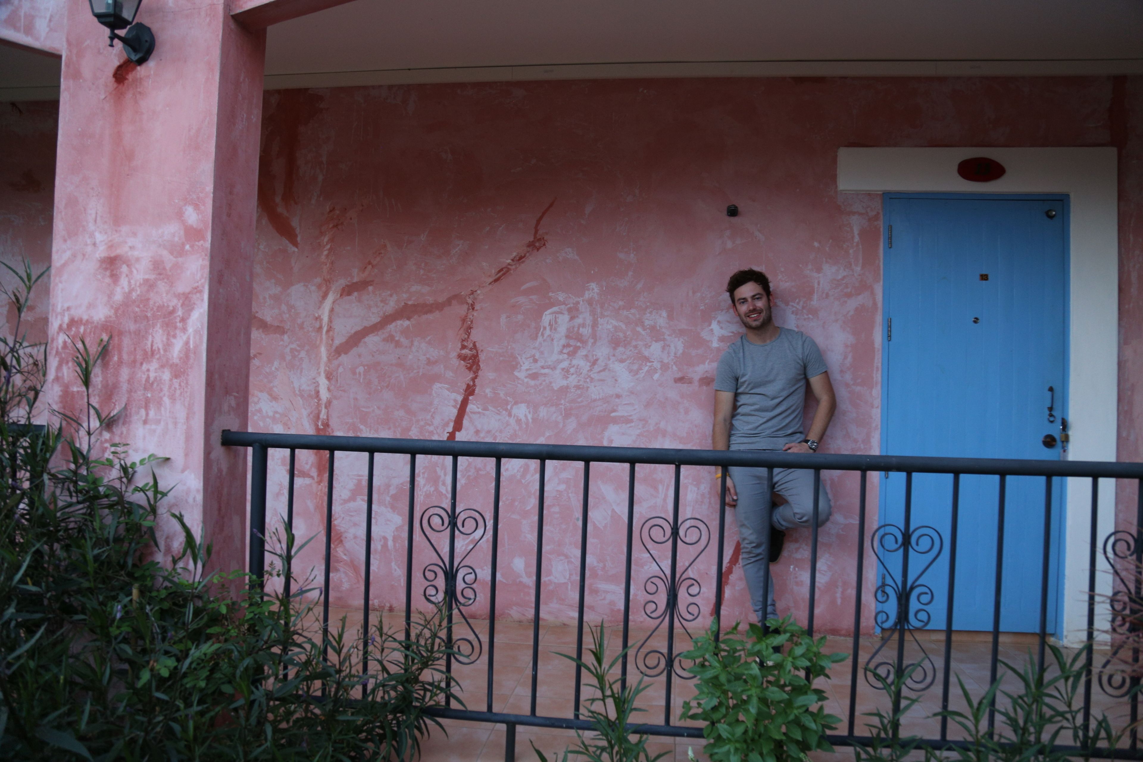 Una persona parada en un balcón con un muro rosa y una puerta azul.