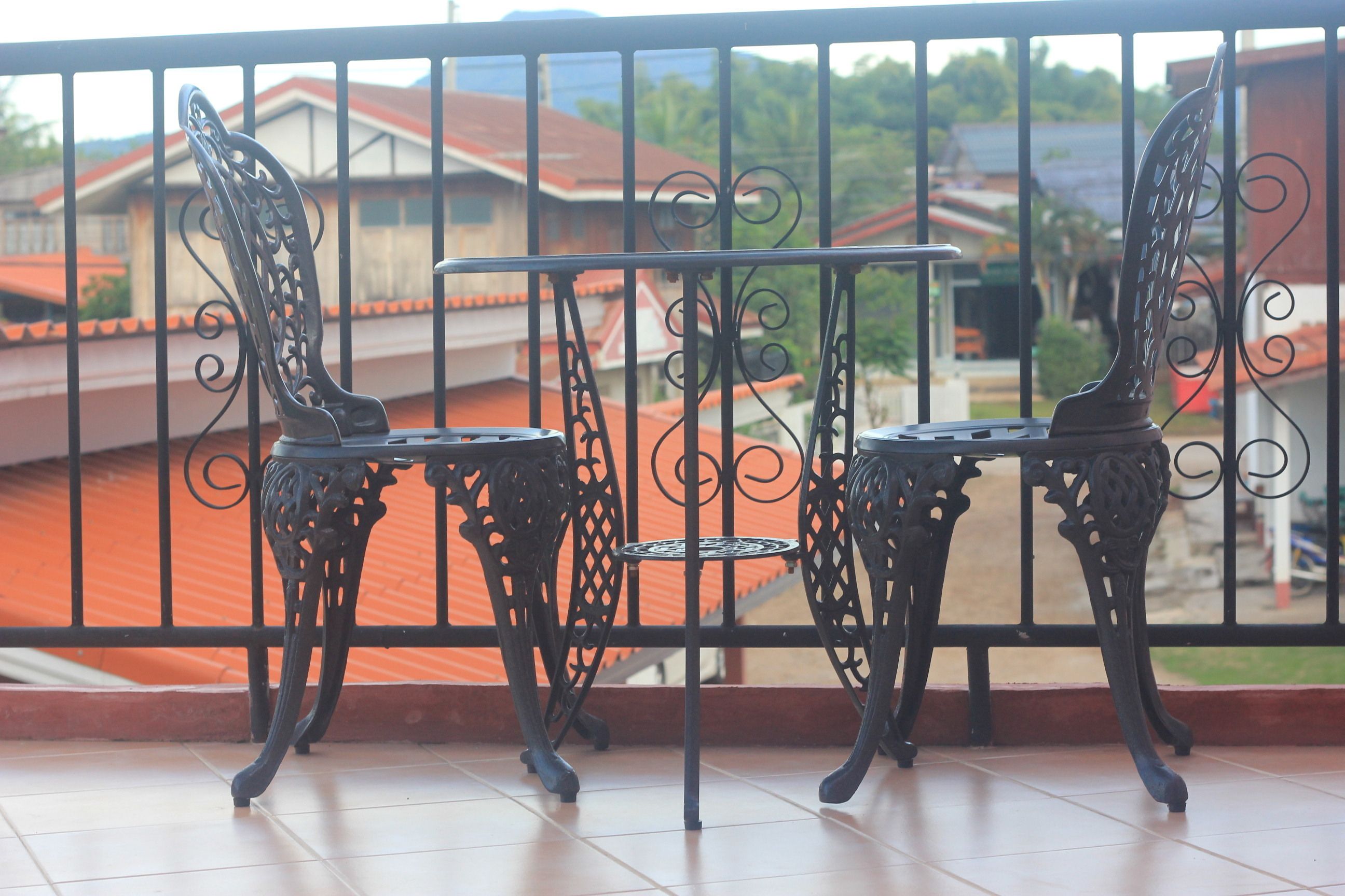 Un balcón con sillas de metal ornamentadas y una pequeña mesa, con vista a una zona residencial de casas con tejados rojos.