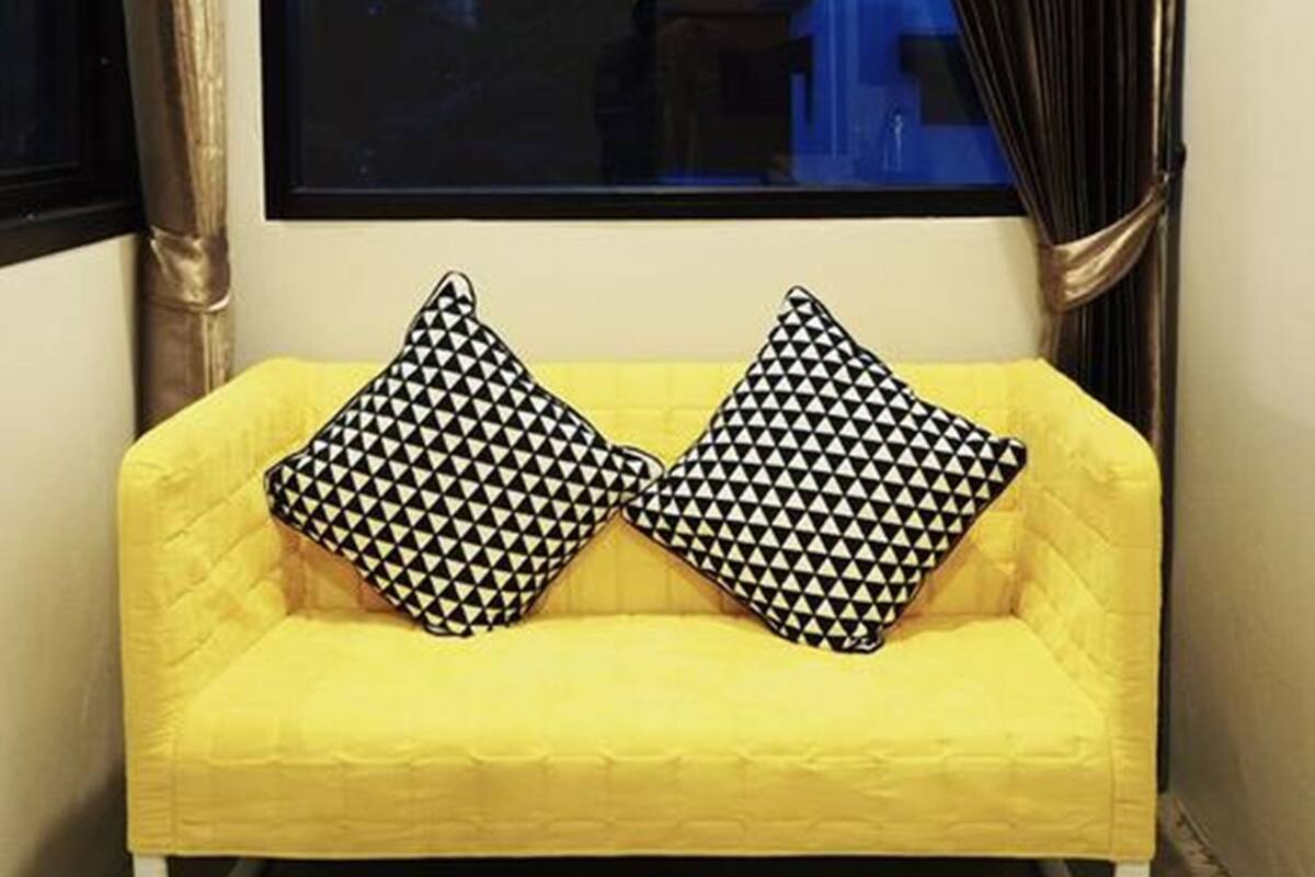 A yellow sofa with black and white patterned cushions, positioned in front of a window with a view of buildings and a blue sky.