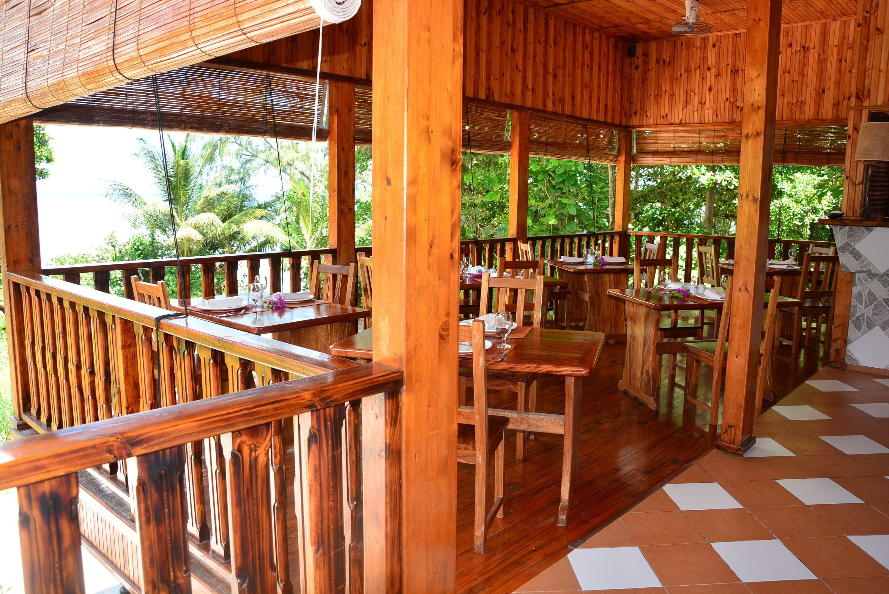 Un área de comedor al aire libre de madera con mesas y sillas, rodeada por una barandilla de madera y con vistas a un paisaje tropical con palmeras.