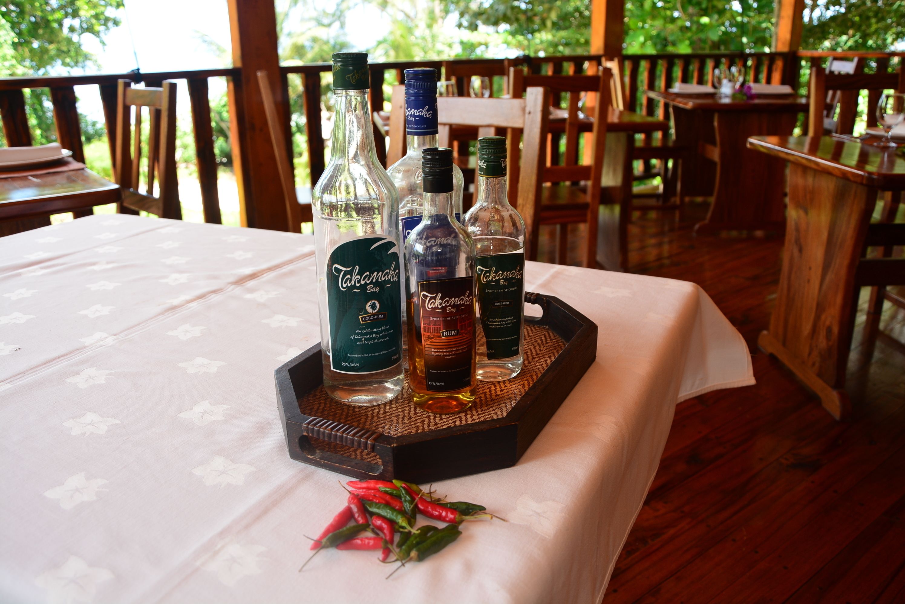 Una mesa con botellas de ron Takomaka Bay y otras licores, una bandeja y algunos chiles sobre un mantel en un área de comedor al aire libre.