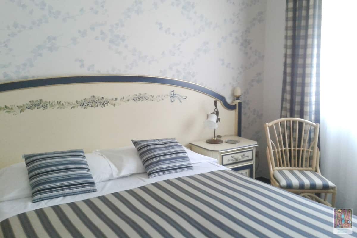 A neatly made bed with striped bedding, a nightstand with a lamp, a chair, and floral wallpaper.
