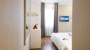 A hotel room with a wooden floor, a bed with white bedding, a TV mounted on the wall, and a mirror on the left wall.