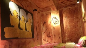 A room with wooden walls, a bed with floral bedding, and a large painting of three figures. - Shannkalay Hostel (Yangon)