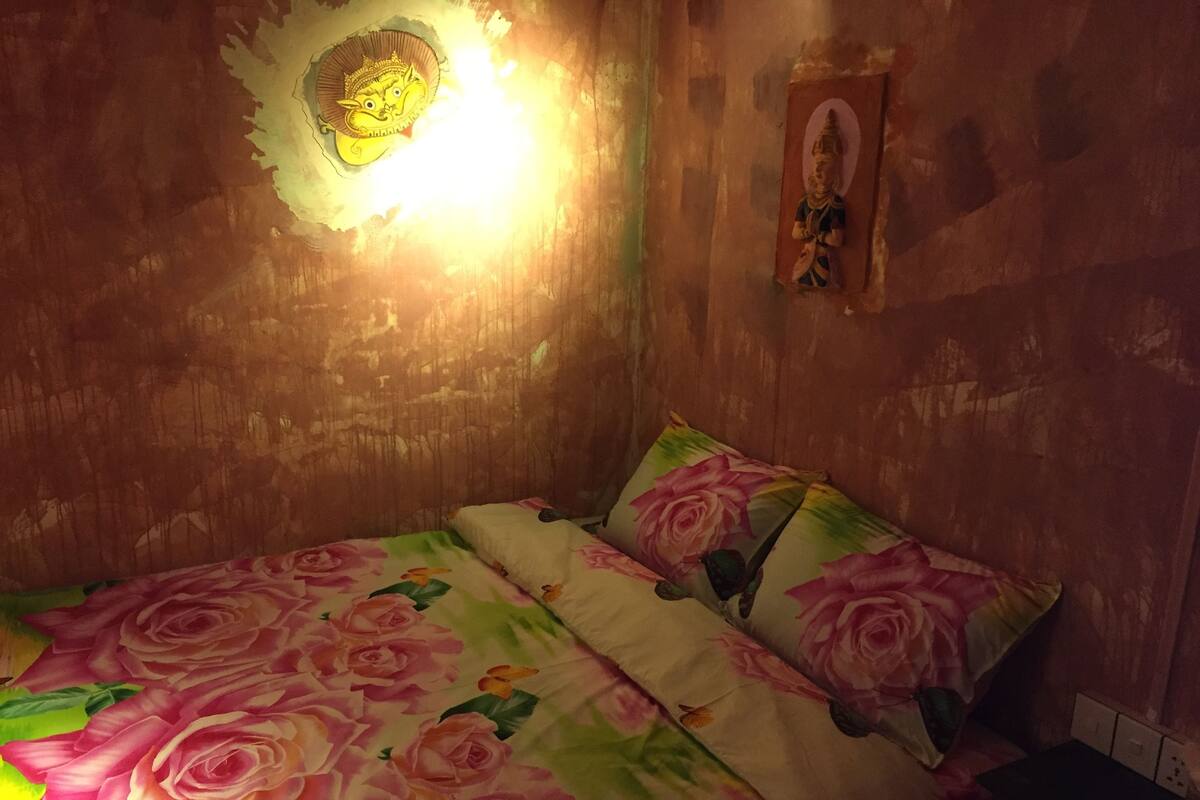 A bedroom with a floral bedspread, a wall-mounted light fixture, and a small religious icon.