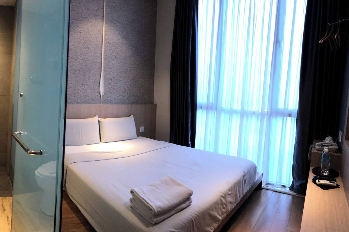 A modern hotel room with a glass shower enclosure, a single bed with white linens, a bedside table with a water bottle, and a view of the outdoors through sheer curtains.