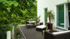 Une terrasse dotée de mobilier d’extérieur, d’une table et de chaises, entourée d’une végétation luxuriante et agrémentée d’un petit élément décoratif aquatique.