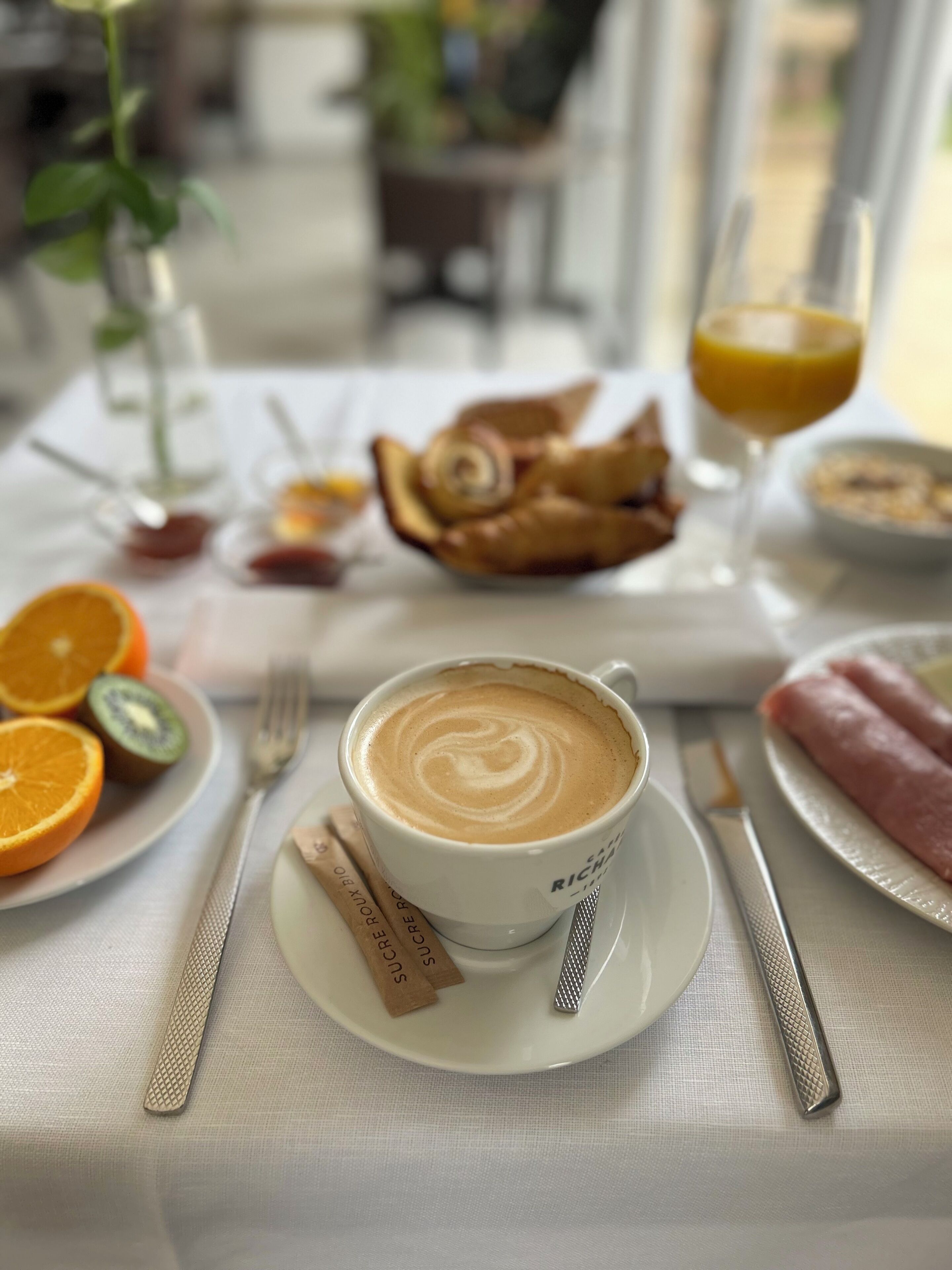 A breakfast setting with a cup of coffee, orange slices, kiwi, a plate of croissants, and a glass of orange juice.