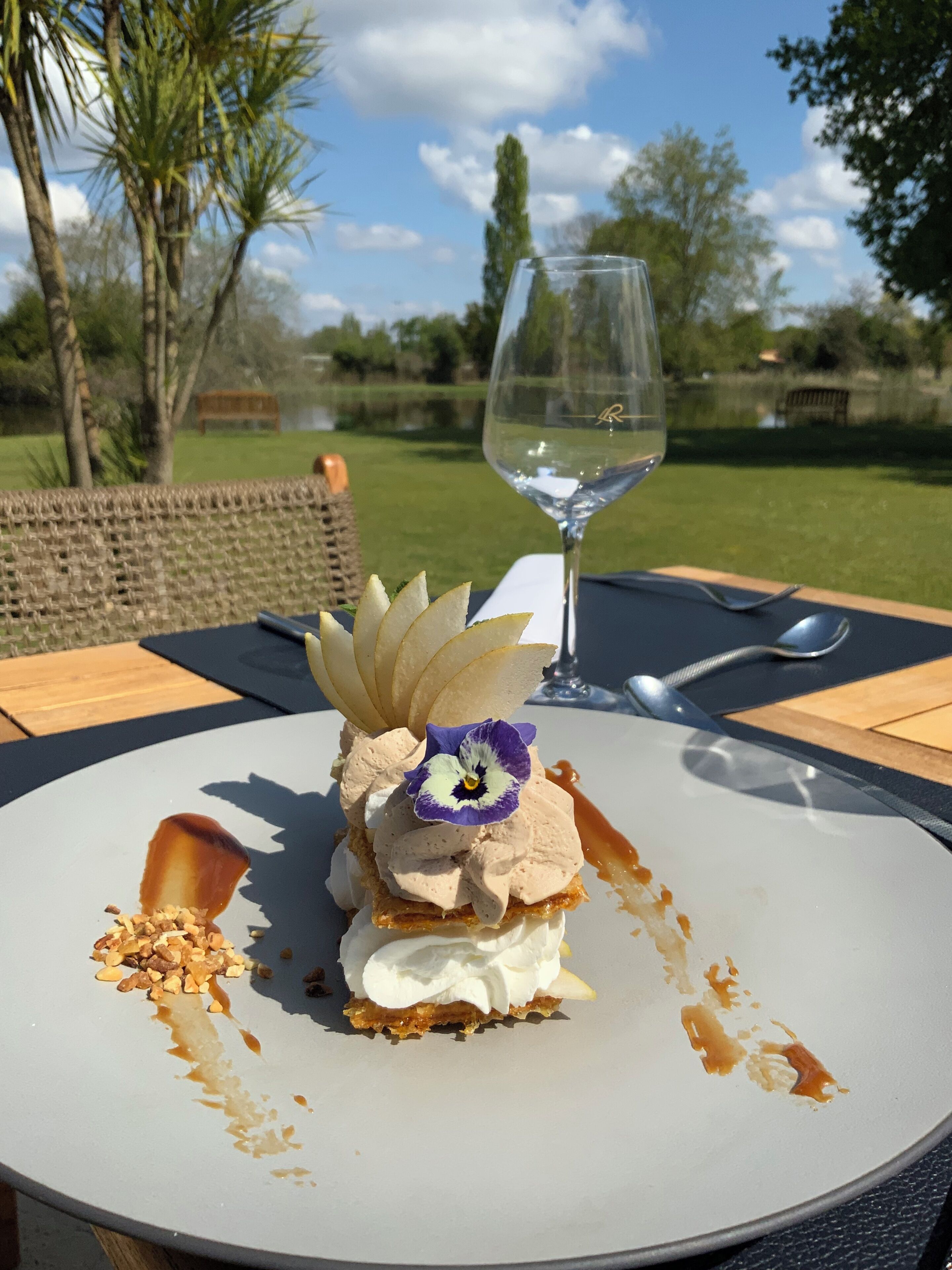 A dessert on a white plate with a glass of white wine on a table outdoors.