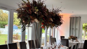 A long dining table set with chairs, a large floral centerpiece, and a view of a swimming pool and greenery outside. - Le Renaissance (Mont-de-Marsan)