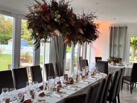 Una larga mesa de comedor dispuesta con sillas, un gran centro de mesa floral y vistas a una piscina y vegetación al exterior.