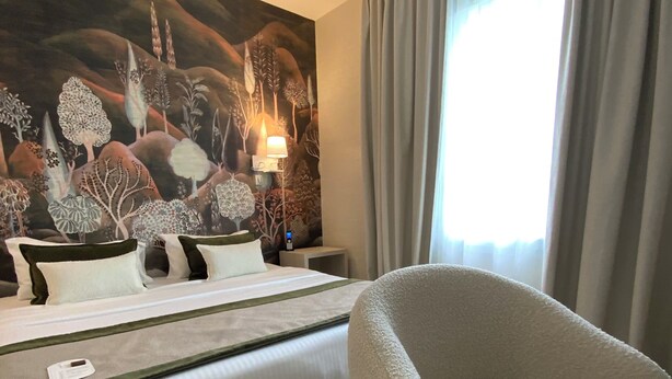 A hotel room with a bed, a chair, a nightstand, a lamp, and a large painting on the wall. - Le Renaissance (Mont-de-Marsan)