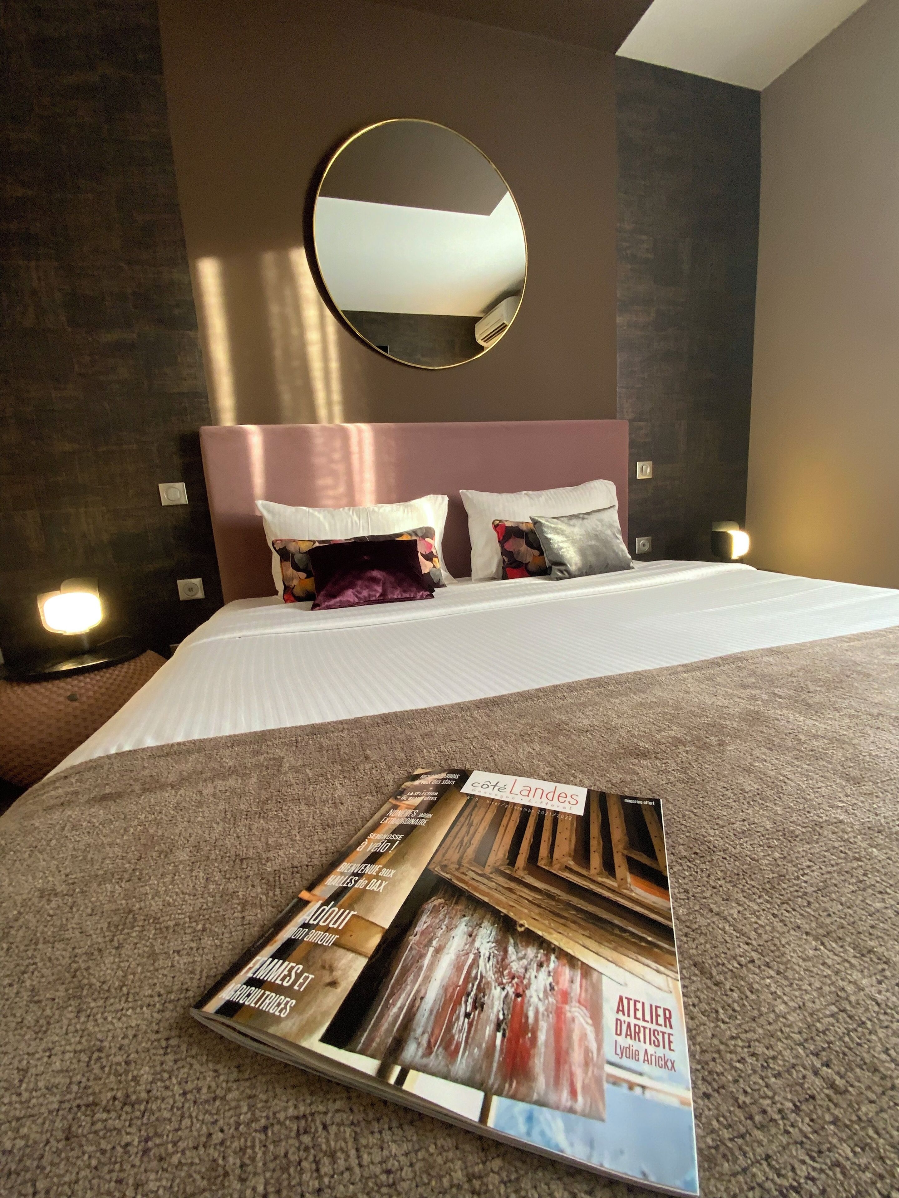 Quarto de hotel com uma cama grande, um espelho redondo acima da cabeceira e uma revista sobre a cama.