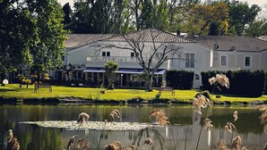 A large white building with a tiled roof, surrounded by a grassy area and a pond with tall grasses. - Le Renaissance (Mont-de-Marsan)