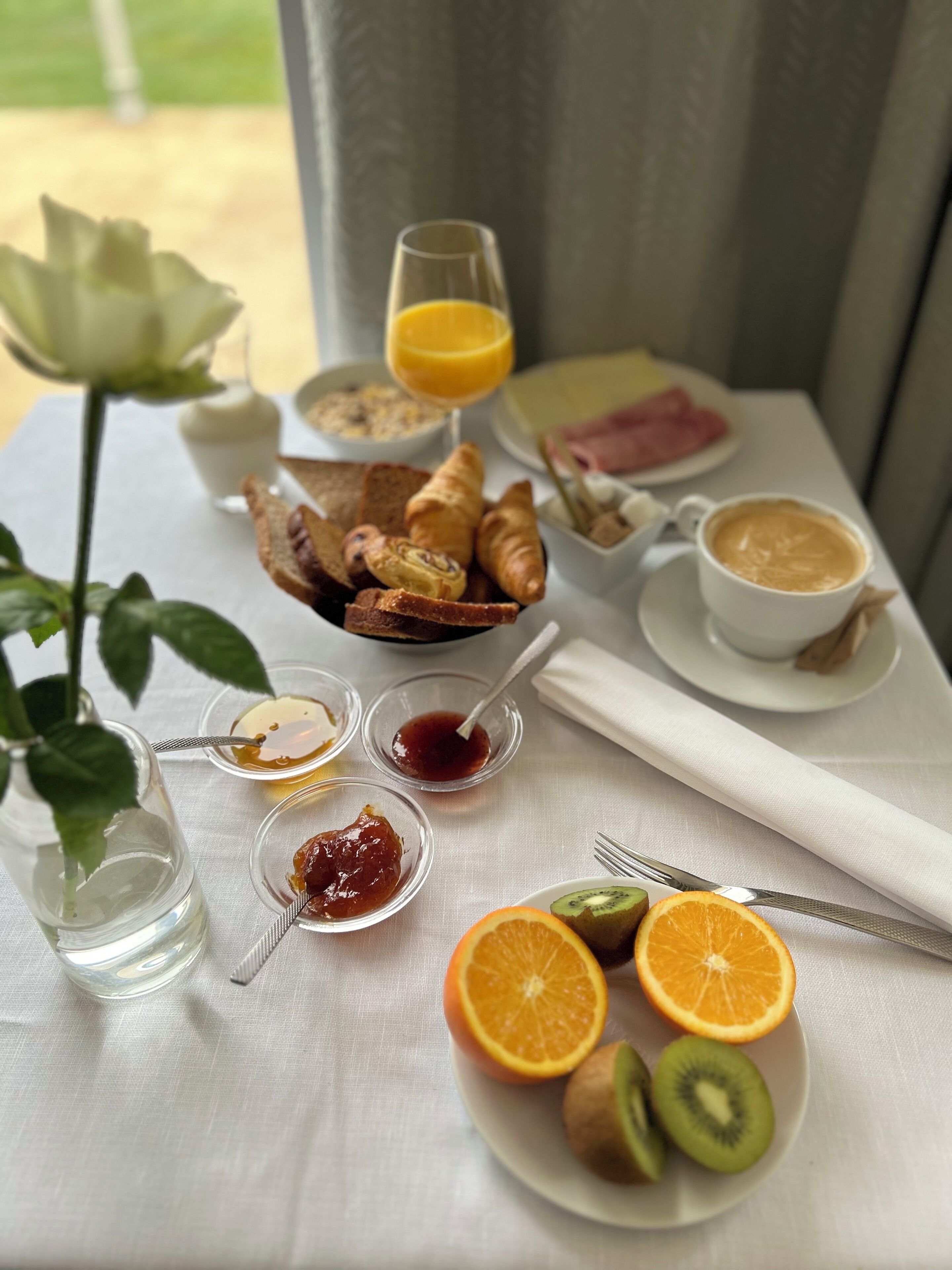 Een ontbijttafel met croissants, toast, jam, sinaasappelschijfjes, kiwi, een roos, een glas sinaasappelsap, een kop koffie en een kleine vaas met een roos.