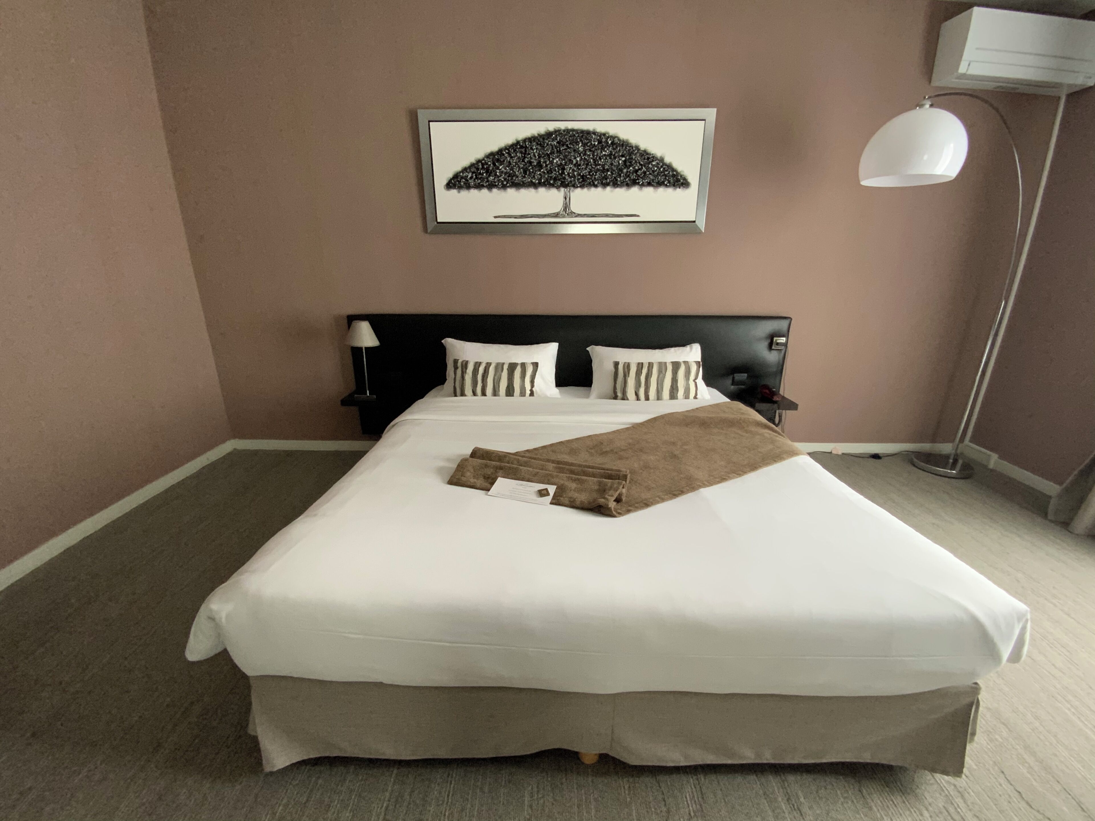 Una cama bien hecha con edredón blanco y manta marrón, cabecera negra y un cuadro colgado sobre la cama.
